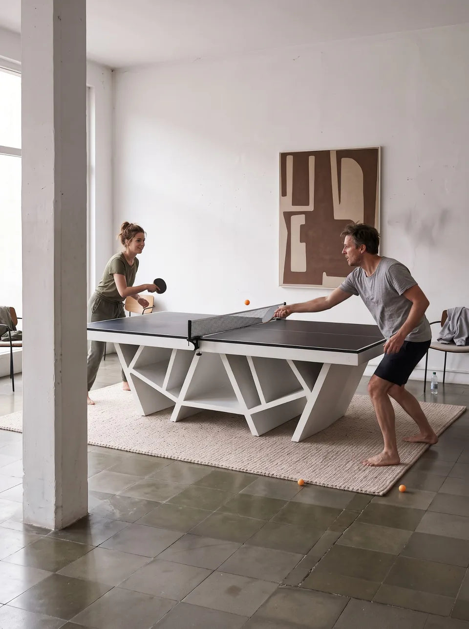 Deux personnes jouant au ping-pong sur une table blanche à piètement en croisillons, colonne de béton et grand tableau abstrait